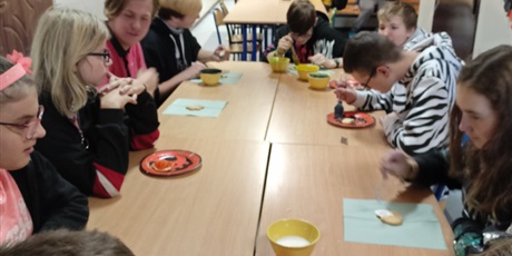Powiększ grafikę: Uczniowie siedzą przy stole i dekorują ciastka kolorowym lukrem.