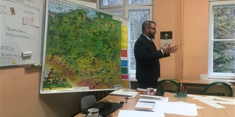 Powiększ grafikę: Pan Arkadiusz Smagacz stoi przy biurku i prowadzi warsztaty, zanim rozwieszona mapa Polski.