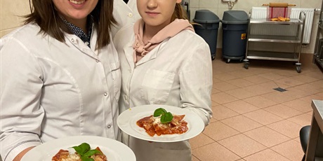 Powiększ grafikę: Uczniowie z nauczycielem stoją w kuchni szkolnej i trzymają talerze ze spagetti.